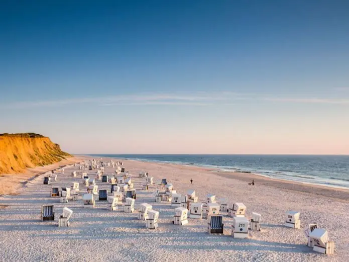 View of Sylt's west beach with long rows of white beach chairs, dunes and the North Sea under a blue sky.