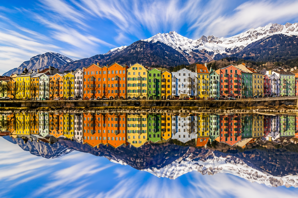 A colorful row of houses on the Inn River in Innsbruck, reflected in the water and with a snow-covered Alpine panorama in the background.